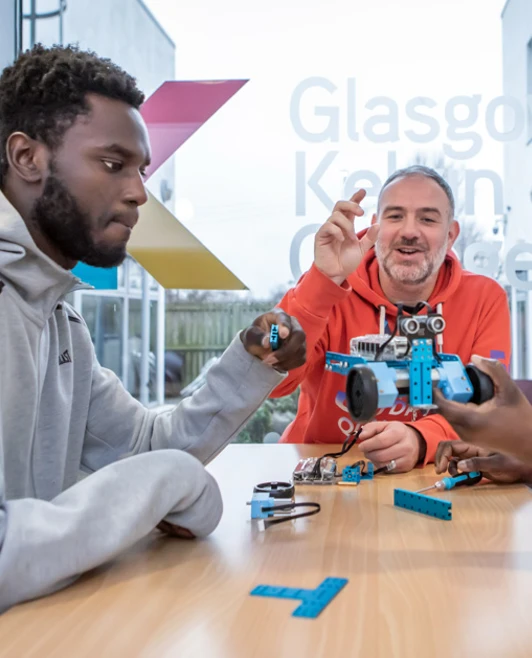 Lecturer in an orange hoodie helping two students troubleshoot and assemble a blue robotic vehicle in a classroom. Lecturer in an orange hoodie helping two students troubleshoot and assemble a blue robotic vehicle in a classroom.
